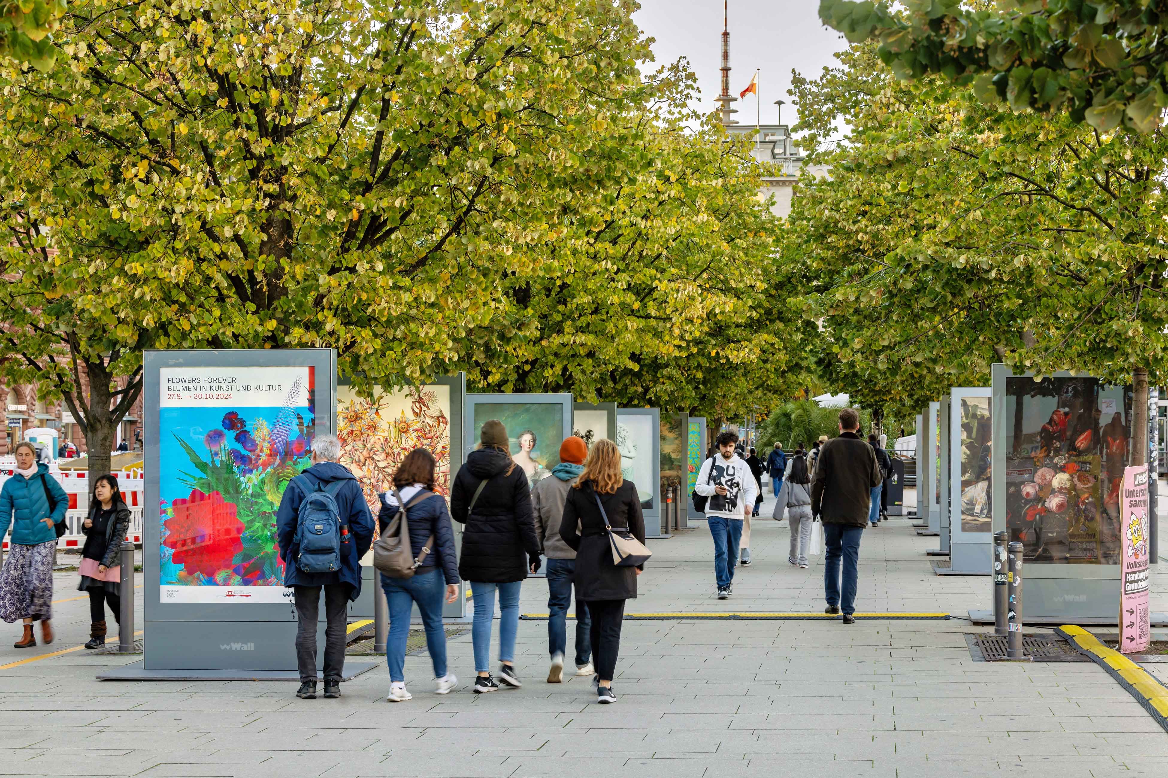 People walk along a city street lined with trees and advertising display boards.
