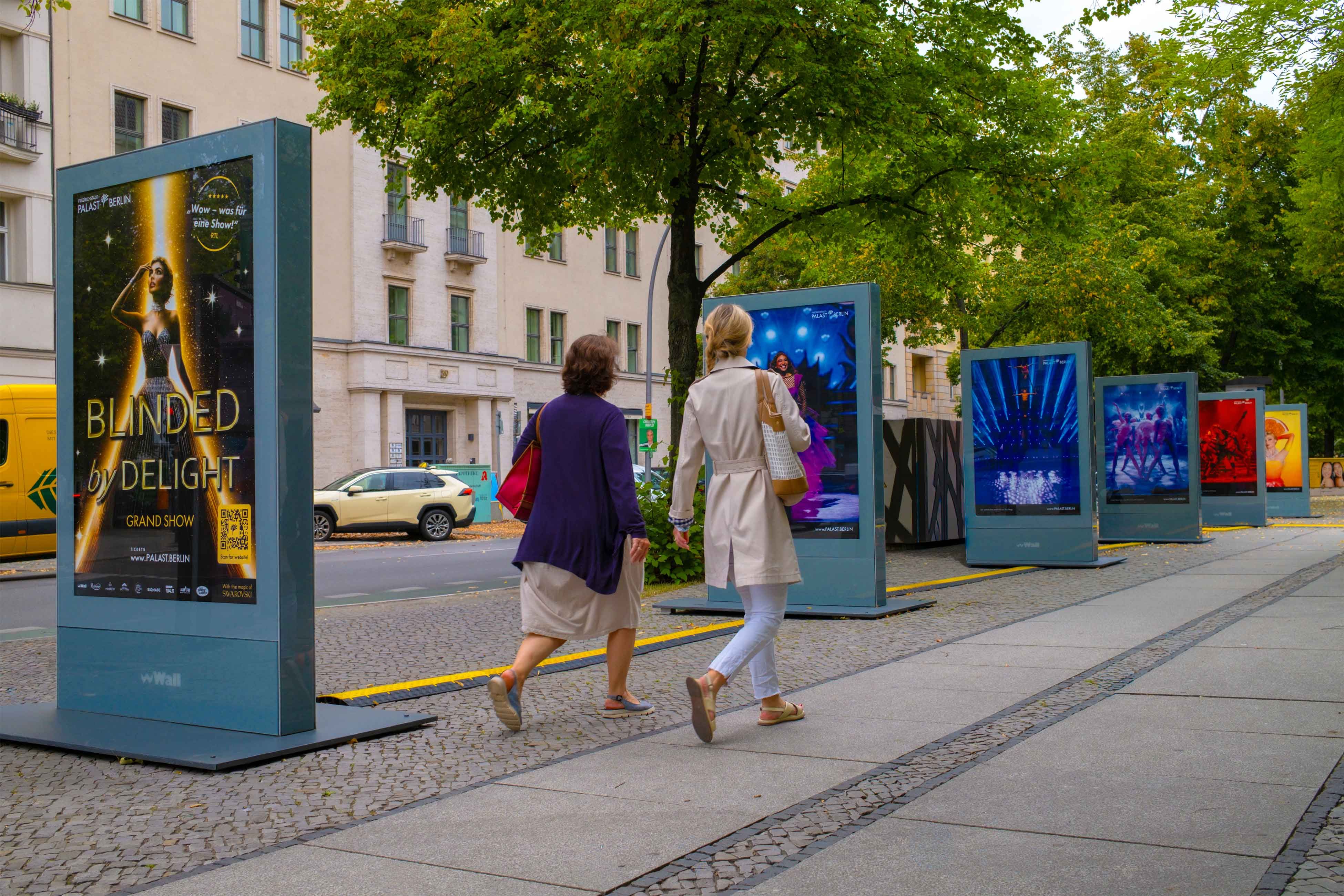 Two women walk past a row of colorful digital billboards on a city sidewalk.